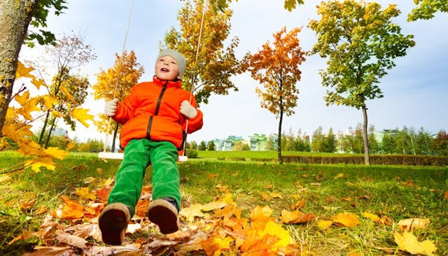 A boy with autism in an orange jacket, green pants, and a gray beanie is smiling and playing on a sw...